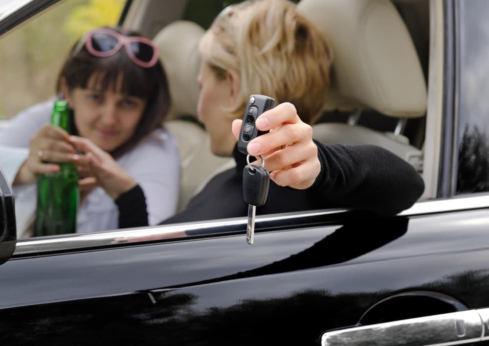 A female driver leans out of her car window, clutching the keys in one hand while reaching for a bottle of alcohol with the other, seemingly cognizant of the danger her drinking poses to fellow motorists.